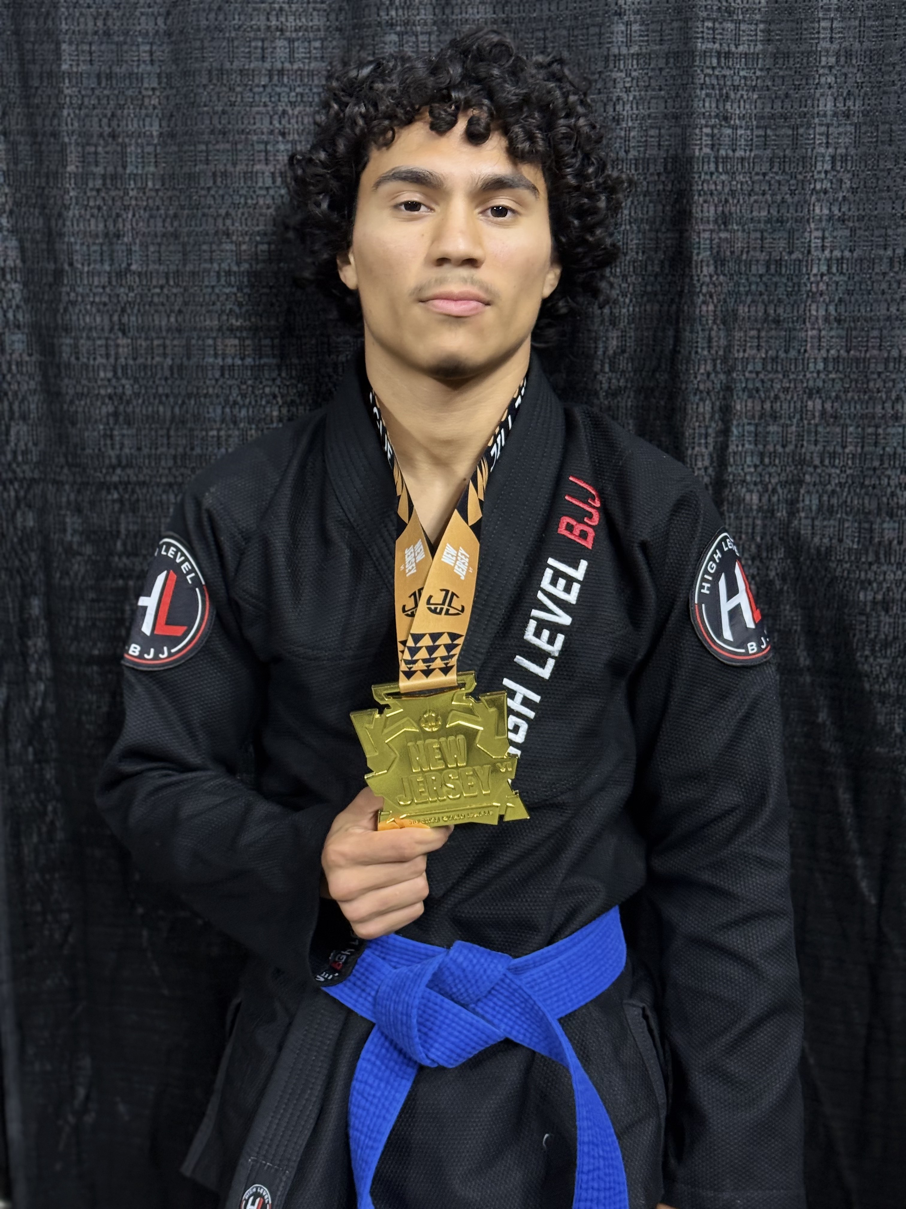 Joel Delorbe in a black gi with a blue belt, holding a gold New Jersey tournament medal.